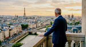Une vue de dos d'un homme d'affaires d'âge mûr, en costume bleu marine et aux cheveux gris, debout sur un balcon en pierre sculpté, regardant un panorama étendu de Paris au coucher du soleil. La Tour Eiffel est visible à gauche, la Seine serpente au centre et Notre-Dame à droite.