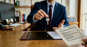Homme en costume bleu assis à un bureau en bois. Il tient une clé d'appartement avec un porte-clés d'appartement dans sa main droite. Sa main gauche est tendue pour recevoir un document papier intitulé "DEVIS DE RÉNOVATION ÉNERGÉTIQUE" des doigts d'une autre personne entrant dans le cadre depuis le bas à droite. Le document montre un logo d'entreprise et un montant. L'arrière-plan montre un bureau bien rangé avec un ordinateur et des étagères remplies
